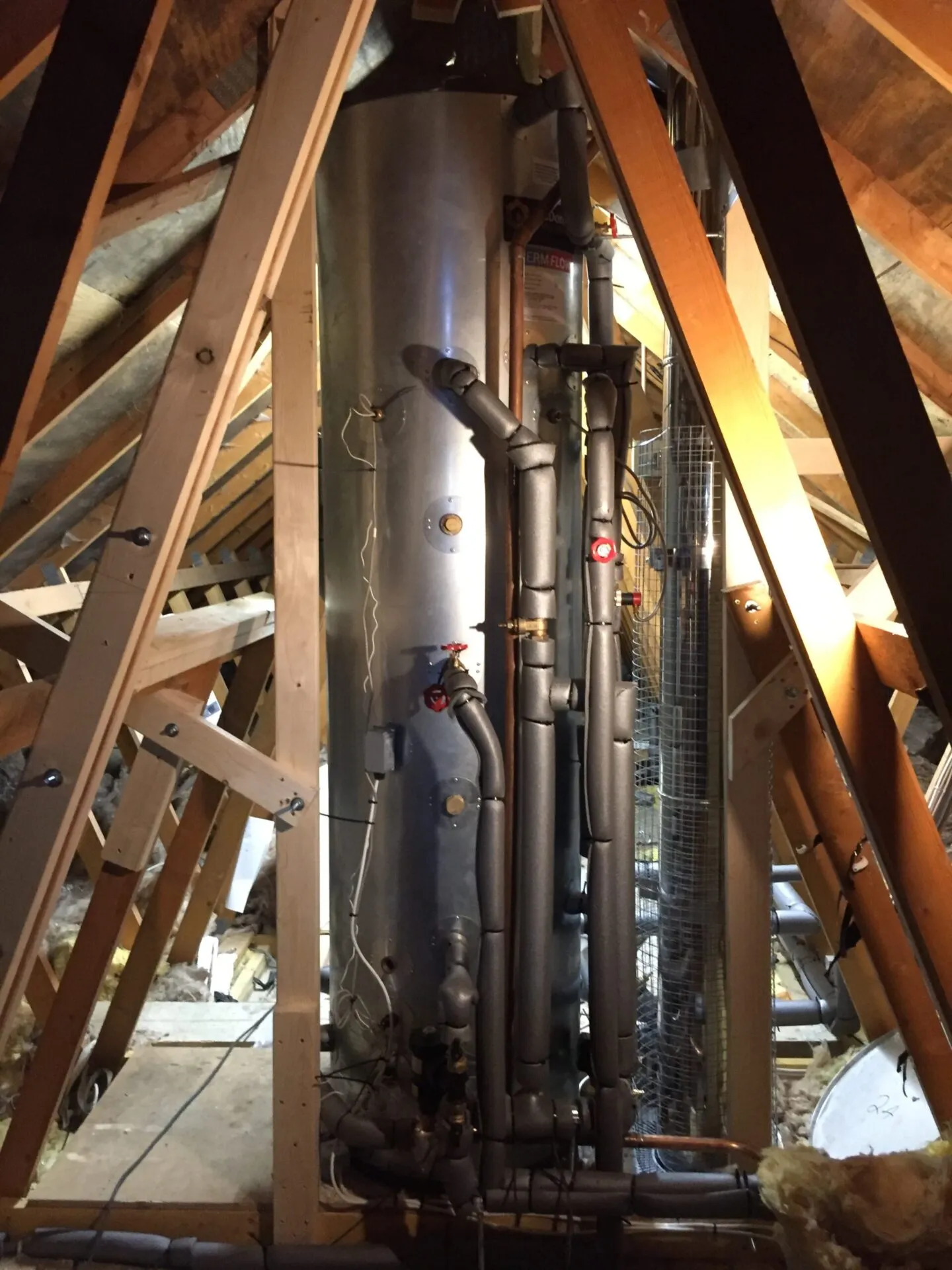 A tall stainless steel thermal store fitted between the roof trusses in a loft, its pipework insulated