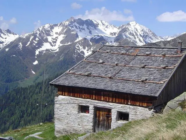 A stone alpine barn with a slate roof and a chimney, snow-capped mountains behind