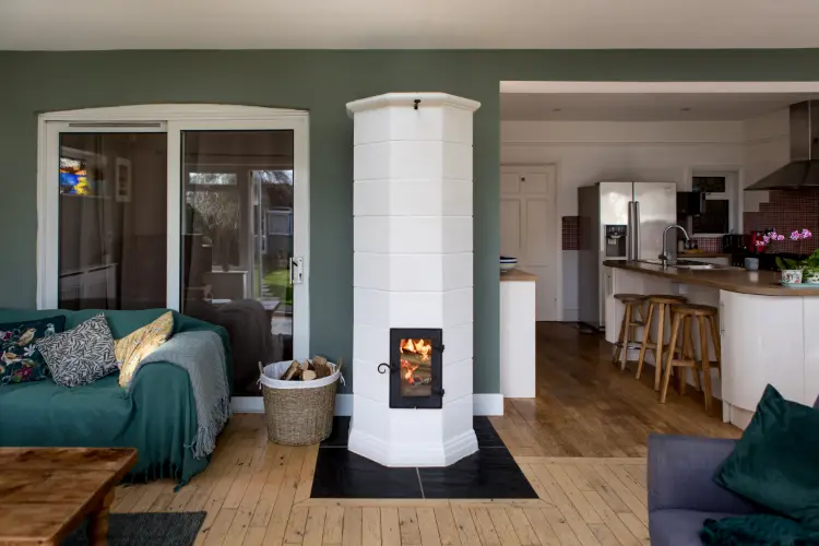A white‑tiled masonry heater alight in an open‑plan kitchen and living room